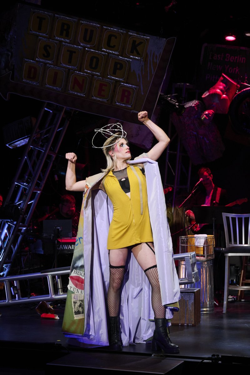 Performer in yellow dress, fishnets, and lavender cape, arms raised, on a stage with a "Truck Stop Diner" sign.