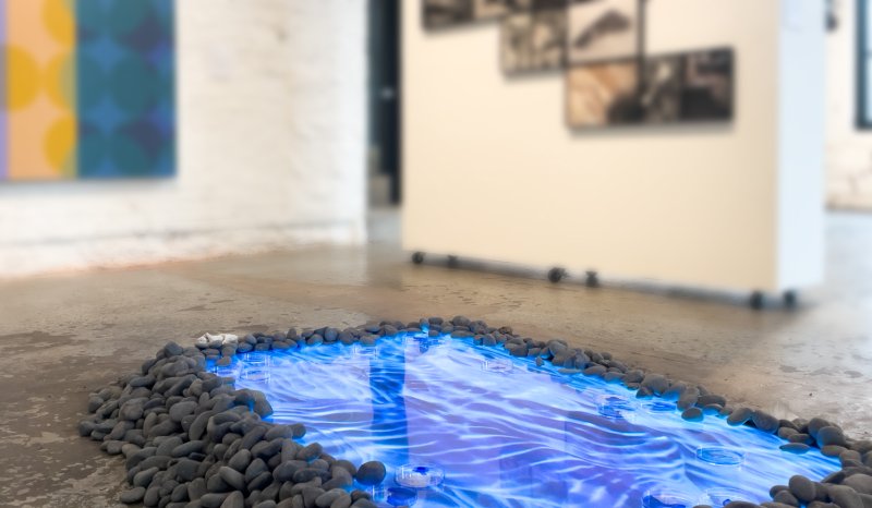 A glowing blue "reflecting pool" art installation on a concrete floor, surrounded by dark stones.