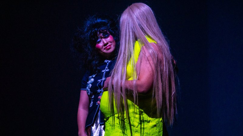 Two figures in dark setting. One with black hair, bright pink blush, and patterned black top. The other has long blonde hair and a textured neon yellow dress.