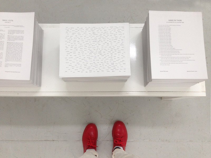 Overhead view of a person's red shoes and white pants standing before three stacks of white paper on a white table.