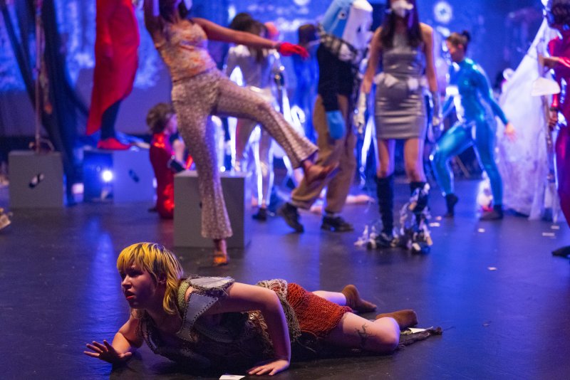 Performance art with a person lying on the floor in the foreground, wearing a brown and green costume. Behind them are blurred figures in costumes of red, silver, and blue, under blue lighting.