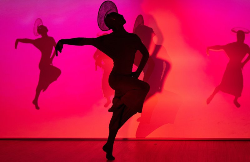 Silhouette of a dancer in a wide-brimmed hat striking a pose, with projected shadows of other dancers on a vibrant pink and red background.