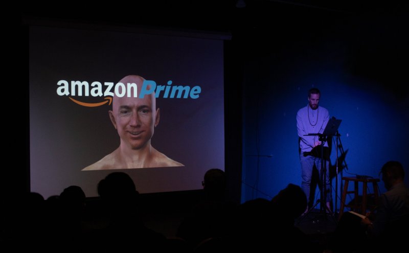 Performance art piece with a projection of "amazon Prime" logo and a bald man's head. A performer stands to the right in blue lighting, with audience silhouettes in the foreground.