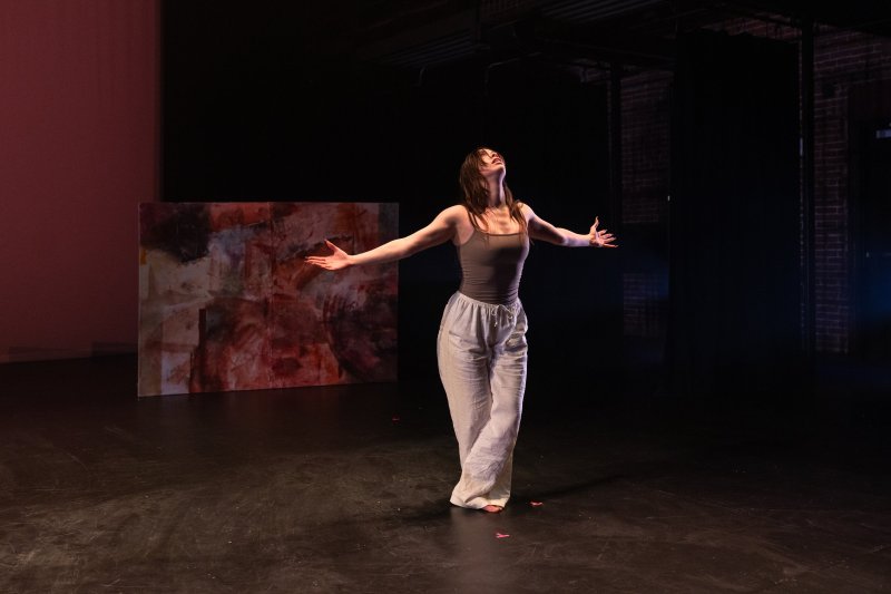 Dancer in a tank top and loose pants, arms outstretched, head tilted back, on a dark stage with abstract red-brown panels.