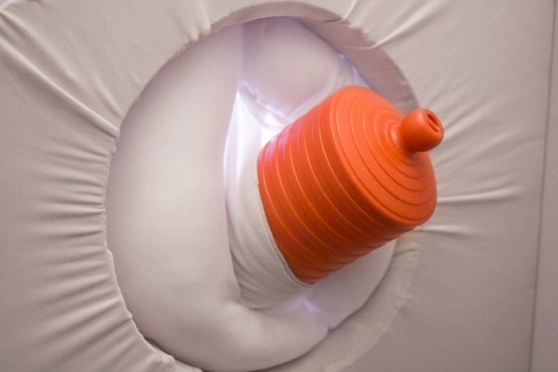 Orange ribbed plunger-like object emerging from a white, soft, fabric-lined opening, with soft white light inside.