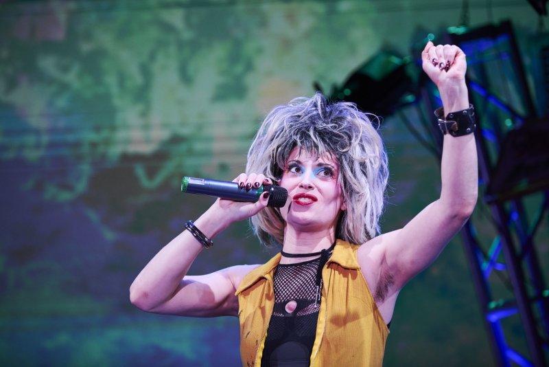 Performer with wild blonde hair, bold blue eyeshadow, red lipstick, black mesh top, and yellow vest, holding a mic with arm raised.