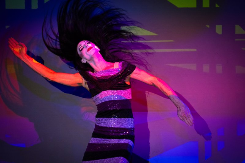 Performer in a sequined dress with long dark hair flung back, arms outstretched, bathed in vibrant pink, purple, and blue stage lights.