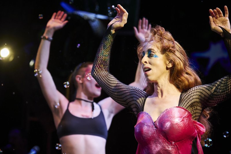 Drag queen with red hair, blue eyeshadow, and a pink corset, arms raised, performing on stage with another person.