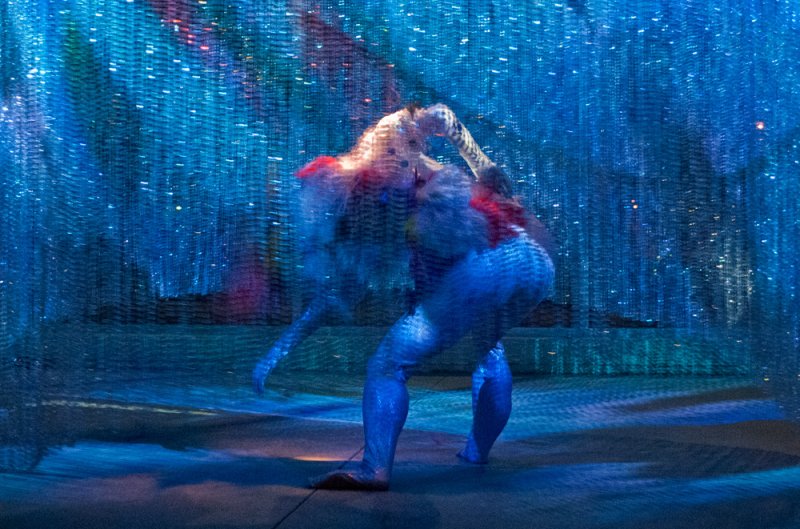 Blue-toned figure with red and white accents, blurred by motion, against a shimmering blue textured background.