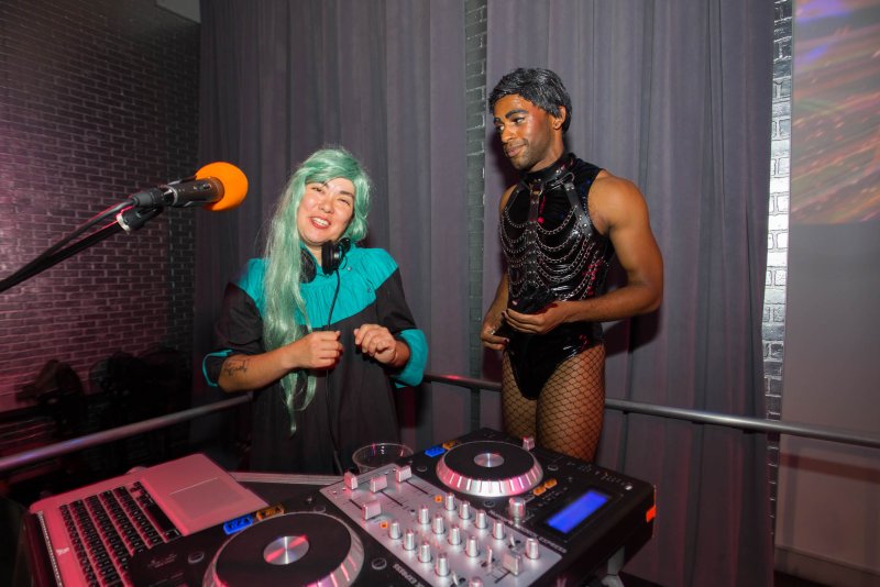 A person with mint green hair DJing, smiling at a person in a black leather harness and fishnet stockings.