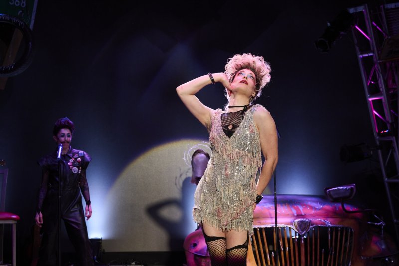 Performer in drag with blonde wig, sequined dress, and fishnets, posing on stage with another performer and car prop.