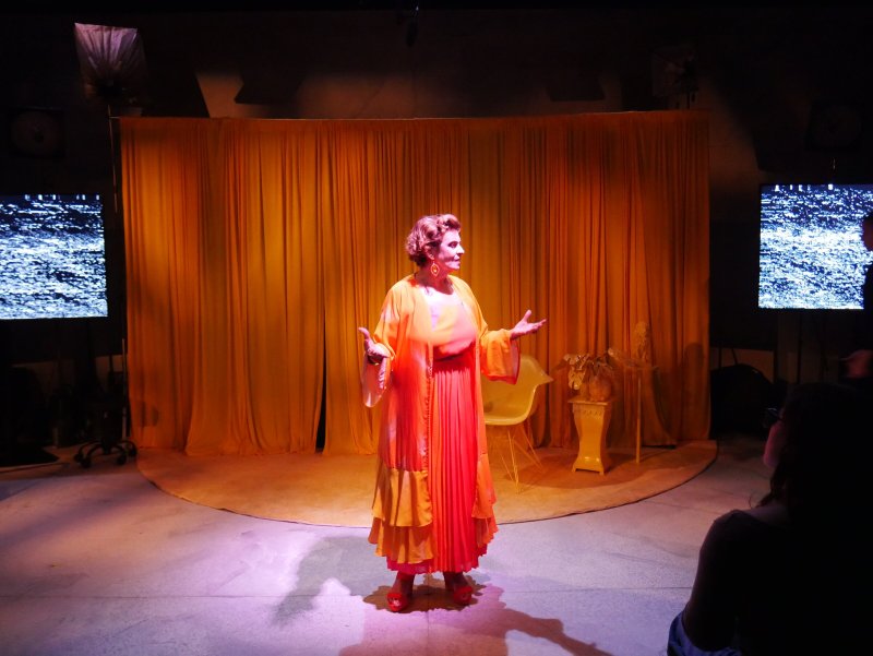 Performer in orange and yellow flowing dress on stage with yellow curtains, two screens with static, and a chair.