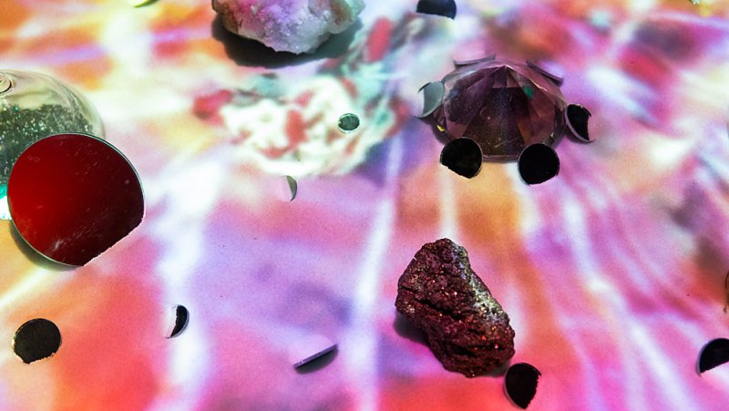 Still life with crystals, a red disc, and black circles on a surface with projected pink, white, and purple light patterns.