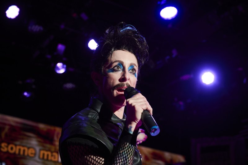 Performer with dark spiked hair, blue eyeshadow, and a mustache sings into a microphone under purple stage lights.