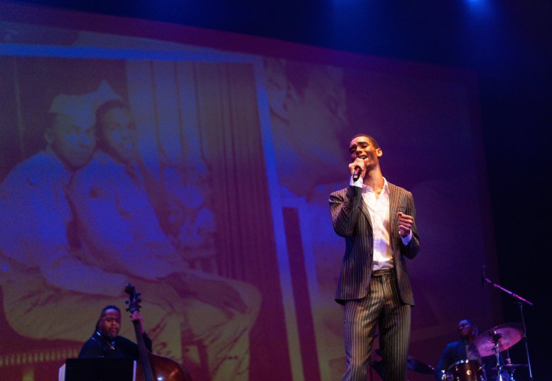 A man in a striped suit sings into a microphone on stage, with a large projected image of two men in uniform behind him.