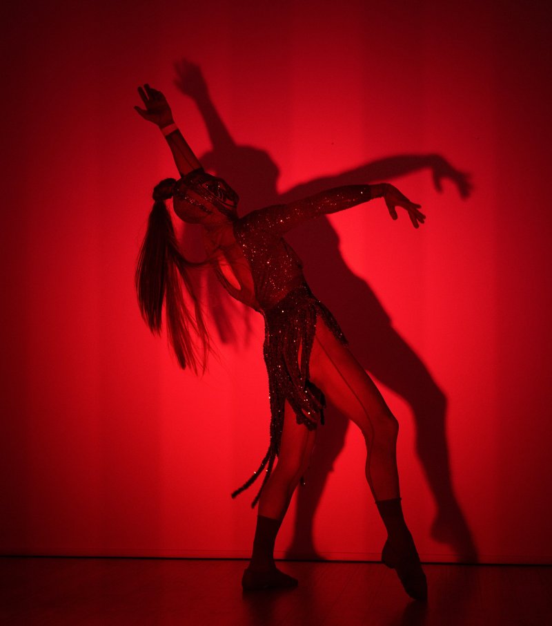Dancer in a sequined costume with long hair, posing dramatically against a bright red background with a strong shadow.
