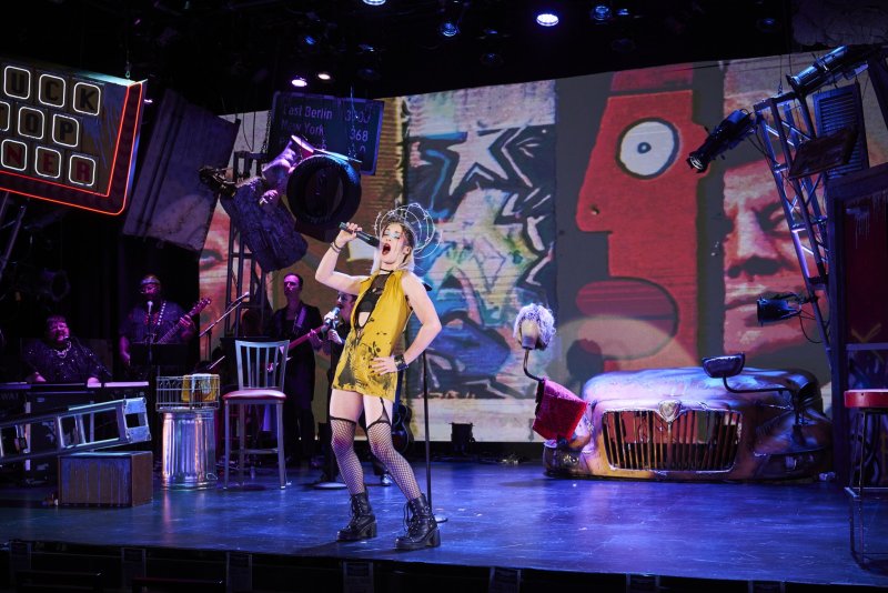 Performer in yellow dress, fishnets, and crown sings into a microphone on a stage with a band, a car prop, and a large, colorful backdrop.