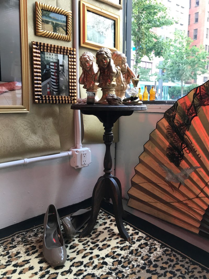 Three golden busts of cherubs on a dark wooden pedestal table, with framed art, a large fan, and shoes nearby.