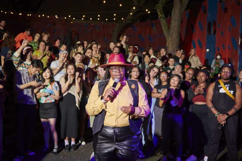 Performance art piece shows a speaker in a red hat and yellow shirt addressing a diverse crowd outdoors at night, with a backdrop of red and blue abstract art and string lights.