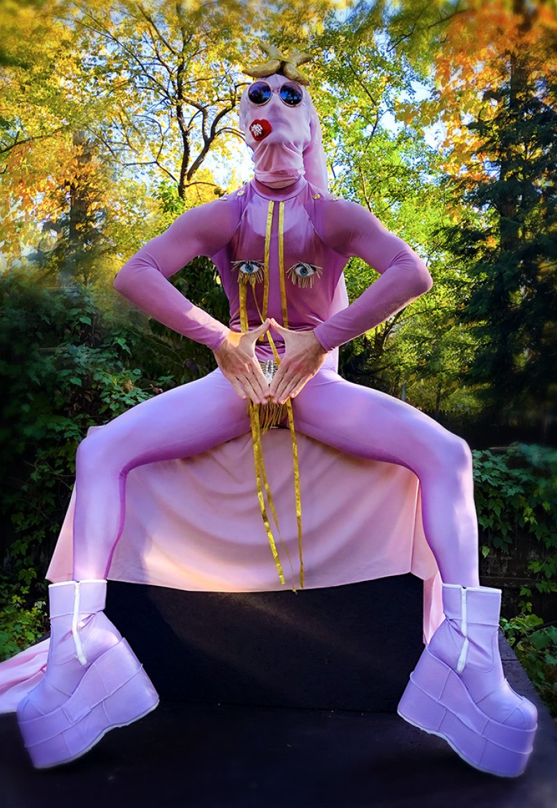Person in full pink bodysuit, platform boots, and sunglasses, with gold tassels and eye motifs, outdoors.
