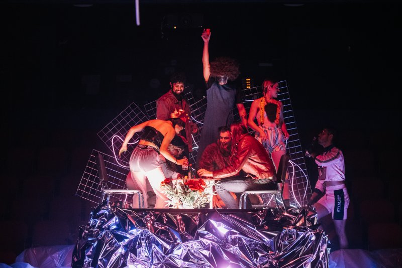 Seven performers on a stage with a crumpled silver table, red roses, and a grid backdrop, lit with purple and red.