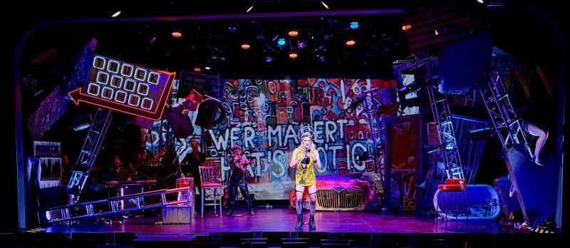 Drag queen in yellow leopard print stands center stage, surrounded by a band, neon signs, and a vibrant, chaotic set.