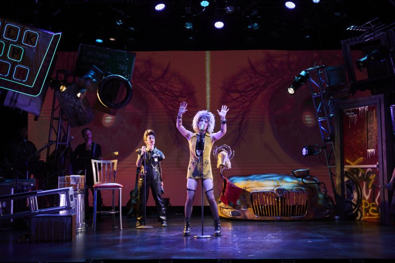 A performer with blonde hair and a yellow dress sings on stage, hands raised, with a band and props in the background.