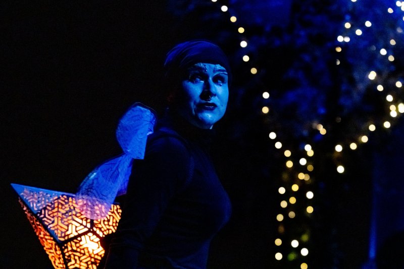 Person in blue face paint, dark clothing, and translucent blue wings holding a glowing geometric lantern. Golden bokeh lights in background.