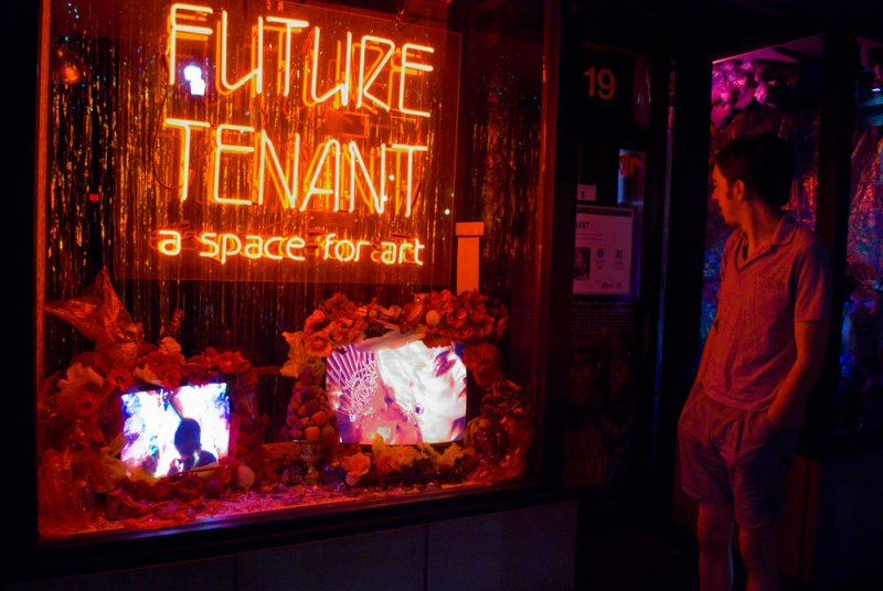 Neon sign reading "FUTURE TENANT a space for art" above two screens surrounded by flowers. A man stands to the right, looking at the installation. Predominantly orange and red lighting.