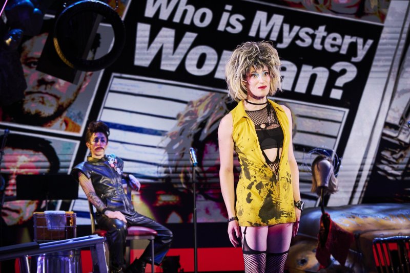Two performers on stage, one in a yellow dress and blonde wig, the other in black leather. A backdrop reads "Who is Mystery Woman?".
