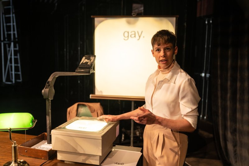 A person with short brown hair stands next to an overhead projector, gesturing towards a screen displaying the word "gay.