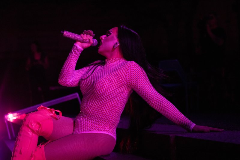 Performer in pink fishnet bodysuit and matching thigh-high boots sings into a glittery microphone under purple stage lights. Dark background.