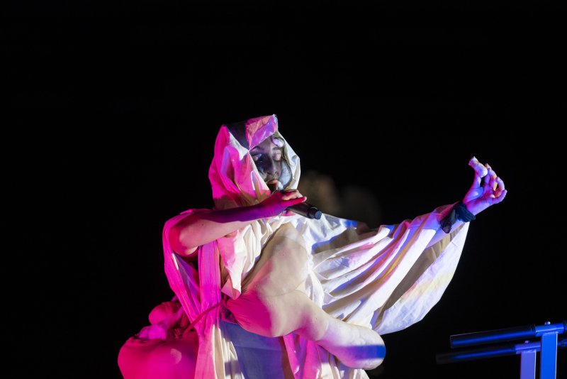 Performer in a hooded, draped costume with warm and cool lighting, holding a microphone, against a dark background.
