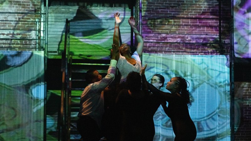 Performance art with five figures against projected brick wall and clock face in blue, green, and purple hues.