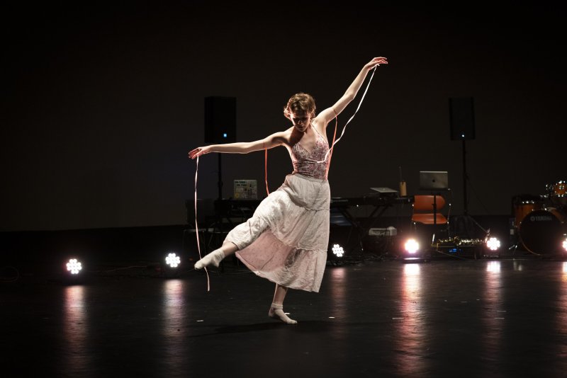 Dancer in a floral top and long white skirt strikes a pose on a dark stage. She holds pink ribbons. Stage lights and musical instruments are visible in the background.