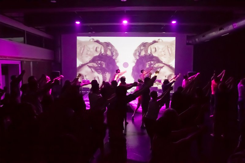 Performance art with silhouetted figures dancing under purple lights, facing a screen displaying mirrored faces.