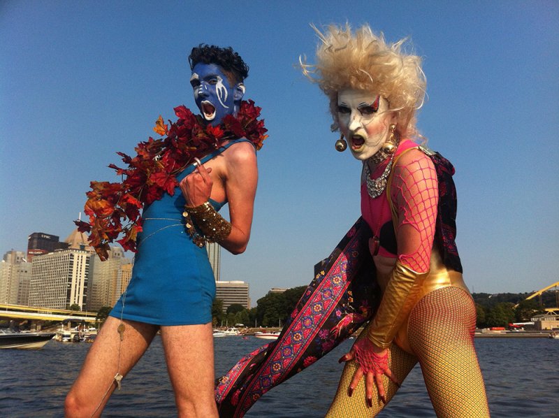 Two drag performers on a boat. One in blue with blue face paint, the other in gold with blonde hair and white face paint.