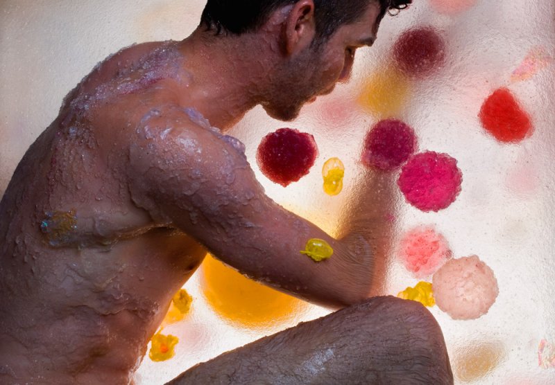 Man with textured skin, covered in clear gel, kneels beside a translucent surface with colorful, organic shapes.