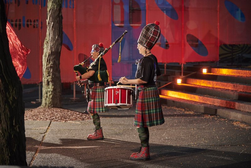 Two figures in kilts and matching plaid hats, one playing bagpipes, the other a red snare drum, stand on a street. Red and blue banners hang behind them, and steps with orange lights are to the right.