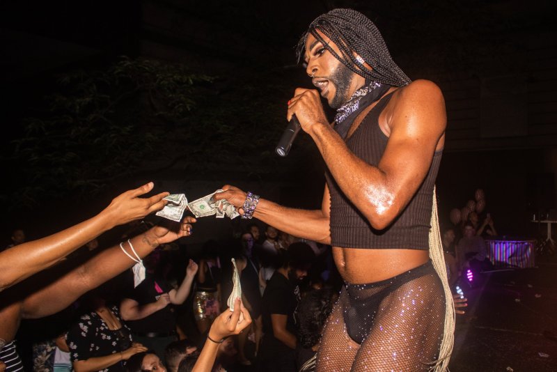 Performer on stage in black crop top and sparkly fishnet tights holds a microphone and accepts dollar bills from outstretched hands in a dark, crowded space.