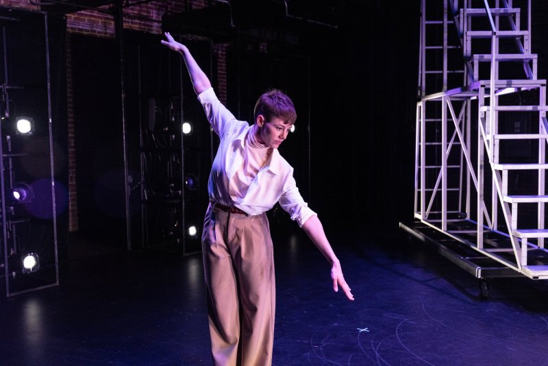 Dancer in white shirt and tan pants on a dark stage, arms extended, with stage lights and metal scaffolding in the background.