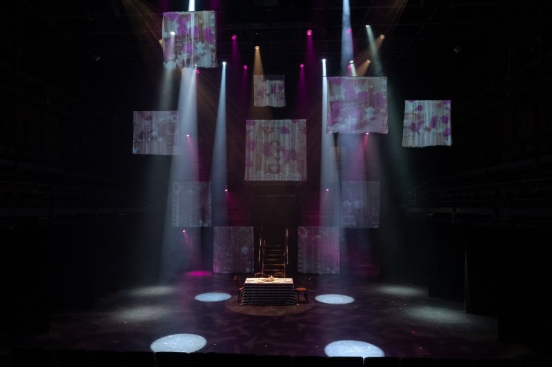 Theater stage with table, chairs, and ladder. Projected floral images hang above, lit by white and pink spotlights.