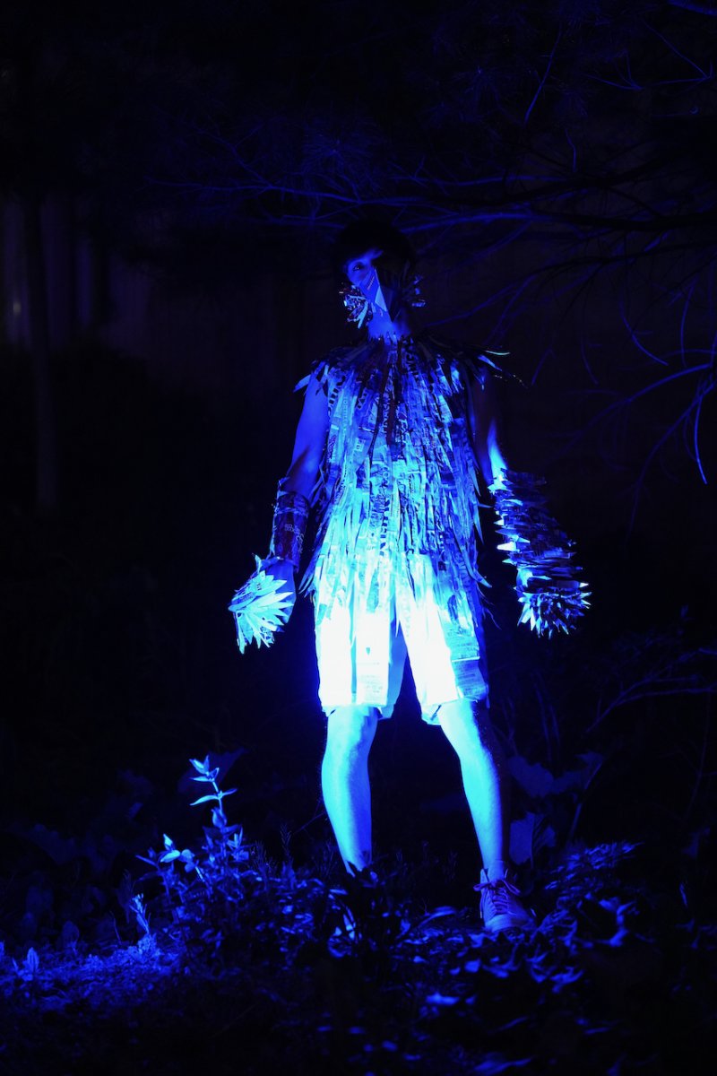 Person in a glowing blue costume made of layered materials, with spiked armbands, standing in a dark, outdoor setting.