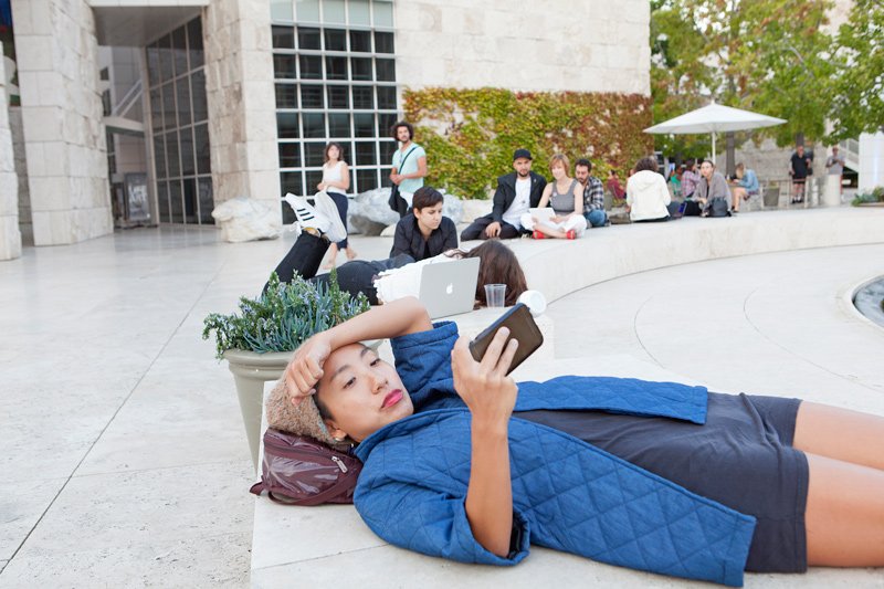 Woman in blue quilted jacket lying on stone ledge, looking at phone, with people and modern building in background.