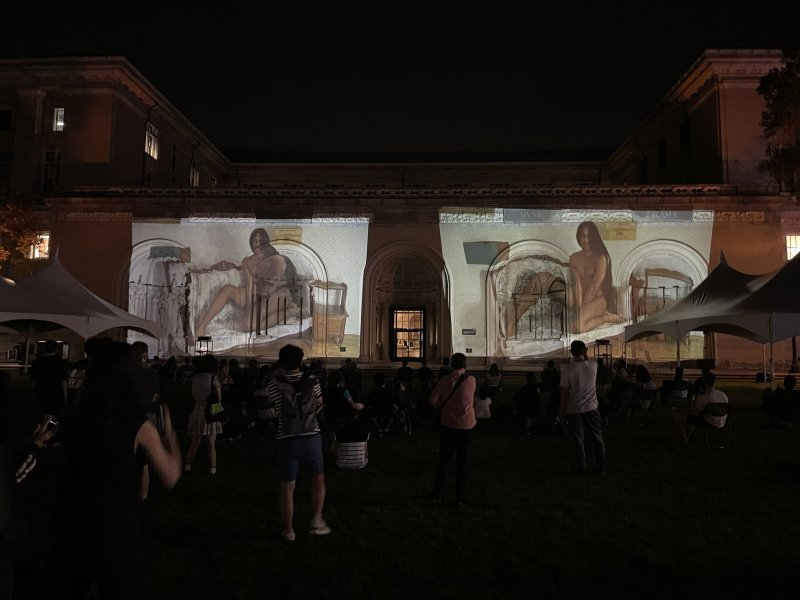 Night view of an outdoor art projection on a building, showing two large, light-colored panels with figures on a dark green lawn with people.