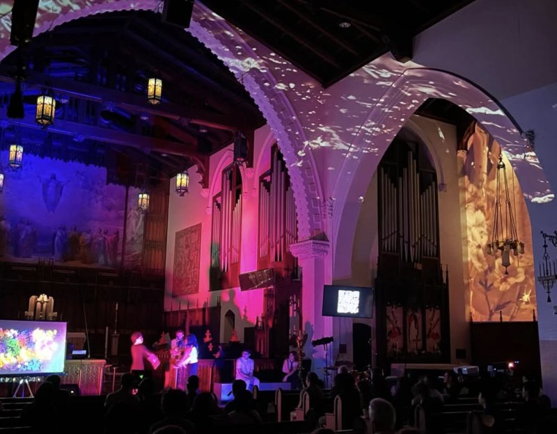 Performance in a church with pink and orange light projections on walls and organ pipes, people on stage, and an audience.