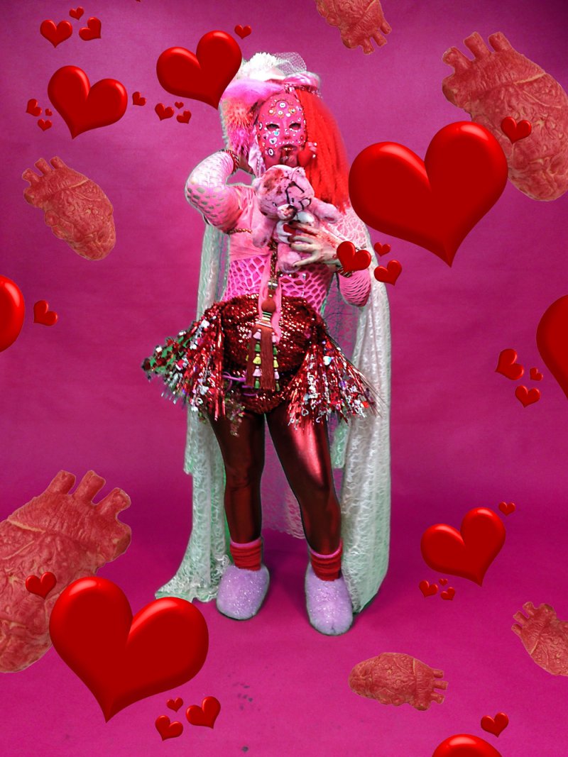 Figure in pink and red costume with mask, holding a teddy bear, against a pink backdrop with floating hearts and anatomical hearts. The figure wears a sequined skirt and a white cape.