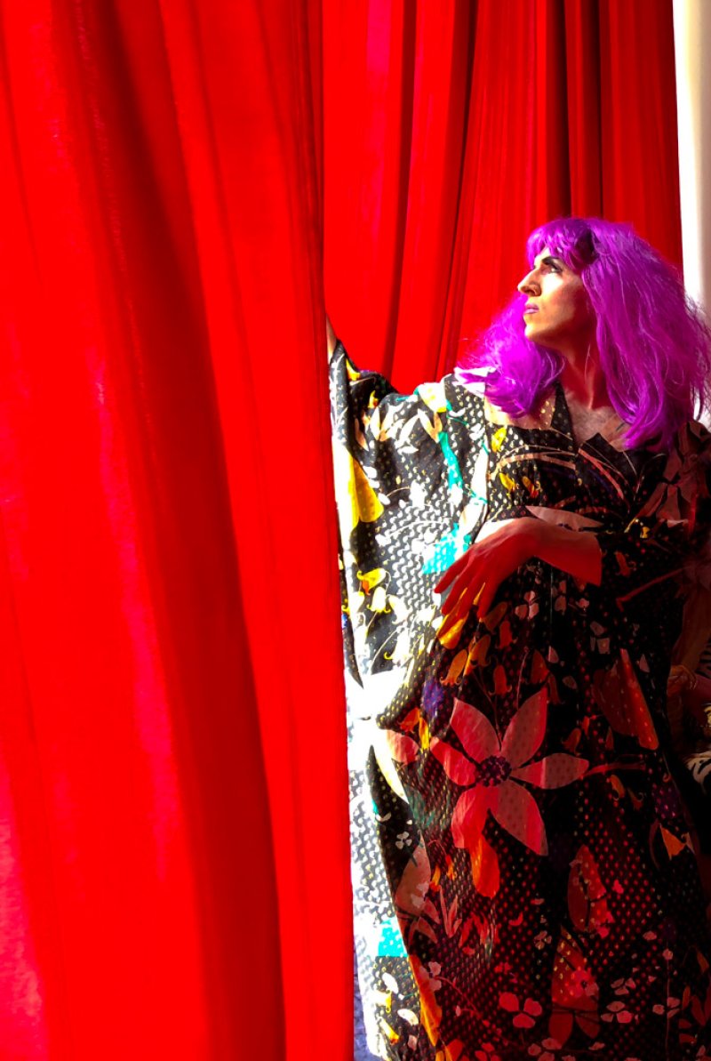 Person with bright purple hair and a floral kimono stands behind a red curtain, looking to the right.