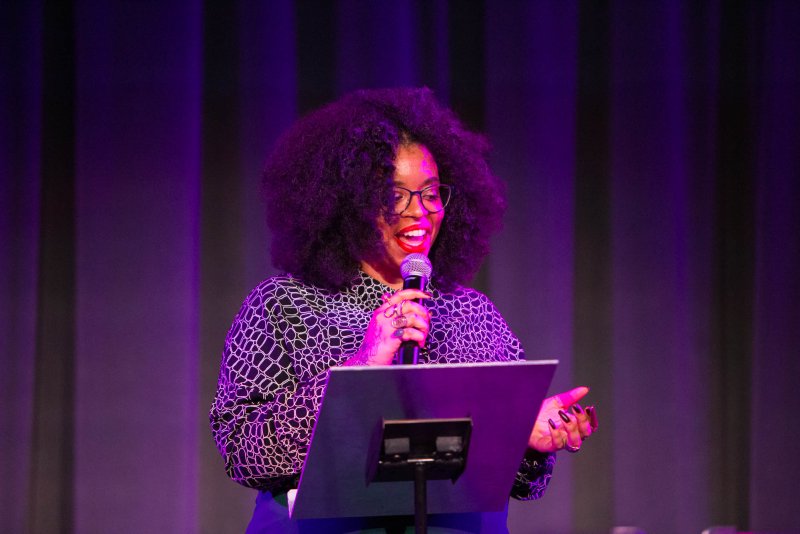 A person with dark curly hair, glasses, and red lipstick speaks into a microphone at a podium, bathed in purple stage light. They wear a patterned shirt and have rings on their fingers.