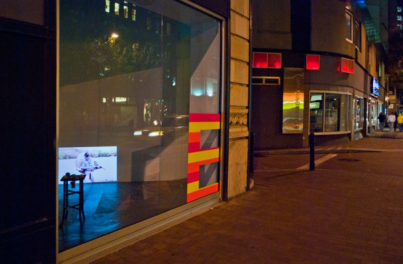 Night street view of storefront window displaying a video on a small screen atop a stool. Window has red and yellow graphic. Street reflection visible.